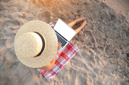 Top view of slender young caucasian woman in hat works on his laptop resting on sandy beach on a sunny day. Slender legs and a hat on a sand background. isolated screen white