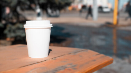 White disposable cup of coffee standing on a wooden table outside, close-up. Street blur in the background, copy space for text