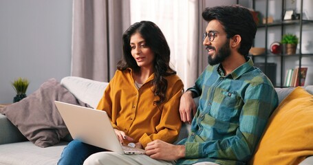 Portrait of young Hindu joyful married couple sitting on sofa in cozy living room browsing online...