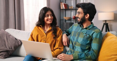 Portrait of young Hindu joyful married couple sitting on sofa in cozy living room browsing online on laptop computer surfing internet choosing something, e-commerce, wife and husband concept