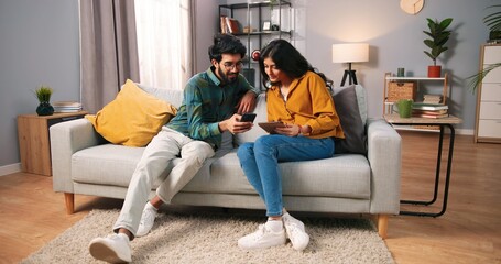 Happy Hindu young married couple resting at home in living modern room using digital gadgets. Handsome bearded husband browsing online on smartphone while his wife surfing internet on tablet