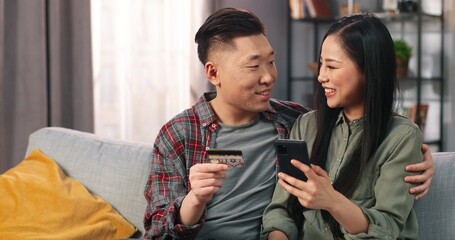 Fototapeta na wymiar Joyful Asian young couple wife and husband sitting on sofa in modern room at home shopping online on smartphone, buying on internet using credit card, making YES gesture, e-commerce concept