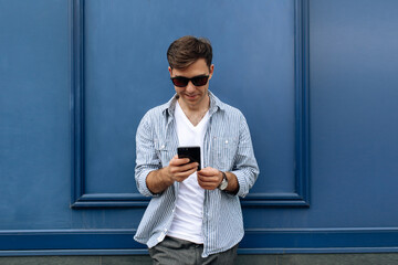 Handsome brunette man in sunglasses smiling while texting on the phone on blue background
