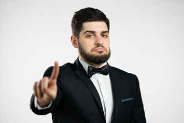Portrait of serious man in black tuxedo showing rejecting gesture by stop finger sign. Guy isolated on white background.