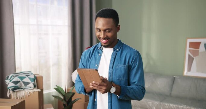 Portrait of cheerful handsome African American young man standing in new apartment smiling and typing on tablet device searching online new furniture and decor for home. Relocation concept, move in