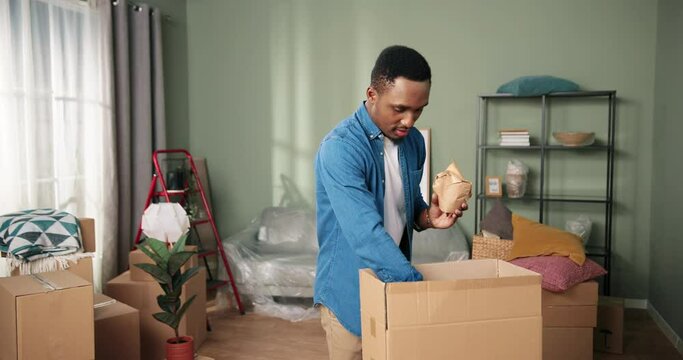 Portrait of African American young handsome cheerful guy unpacking carton boxes decorating new apartment on moving day. Happy man moves in and unpack stuff and furniture. Relocation concept