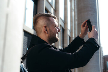 Cheerful cute guy in a black coat makes photos on the phone while walking around the city