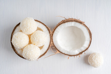 Top view of coconut truffles or homemade vegetarian energy balls with a filling of sweetened shredded coconut and curd cheese served in halved fresh coconut on white wooden table. Horizontal image
