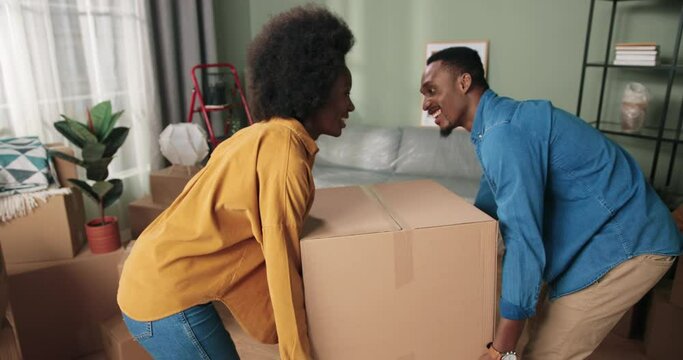 Close up of happy young cute African American wife and husband holding together big carton box parcel moving in new own home. Unpacking stuff in rented apartment. Relocation, removal concept