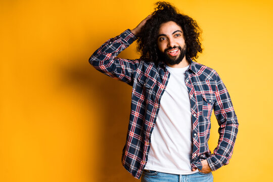 Cheerful young egyptian with a beard in a white t-shirt and shirt holds his head with his hand and stands on a yellow background