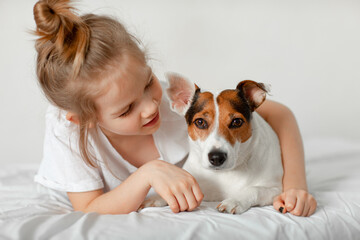 Beautiful funny kid girl lies in bed with a dog in a white bedroom.