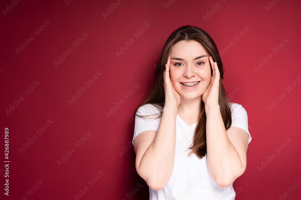 Wall mural a beautiful girl in a white t-shirt with long hair puts her hands to her face and smiles broadly in 