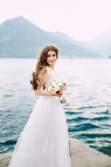 Fototapeta na wymiar The bride holds a bouquet of roses in her hands and stands on the pier in the Bay of Kotor
