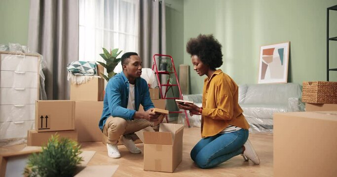 African American beautiful young cute family couple unpacking their stuff together in new apartment on moving day. Happy wife and husband open boxes parcels in own home after renovation Tenant concept