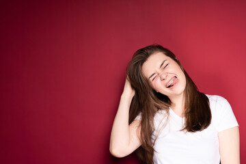 Cheerful young girl stands on a red background and laughs closing her eyes with braces tilting her head to the side