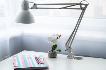 A simple white desk in a homeworker. There is a white lamp on the table. There is a large notebook on the table. The pens are on the notebook.
