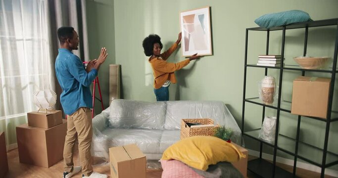 Cheerful young cute married family man and woman move in hanging picture on wall. African American handsome husband saying his wife where to put painting. decorating house, unpacking stuff