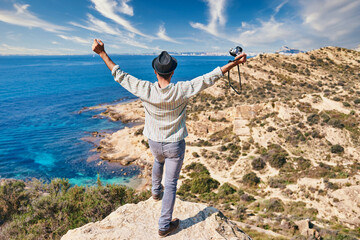 A young man with a hat raises his arms in front of the Mediterranean coast