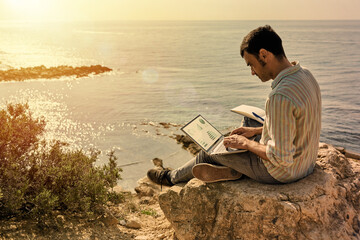 A young man works remotely with the sea in the background