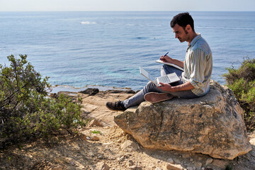 A young man works remotely with the sea in the background