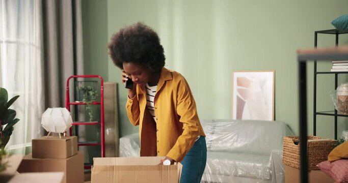 Beautiful cheerful young woman standing in living room new own home talking on smartphone while unpacking big carton box picking stuff and decor. Moving day. Renovation concept. Relocation