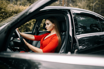 A beautiful young girl in a red overalls sits behind the wheel of a black car on an empty road in the forest