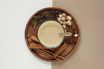 top view of masala chai or Indian spices tea in transparent glass with blurred vision of allspices on brown circle wooden tray placed on cream colour paper and brown paper.
