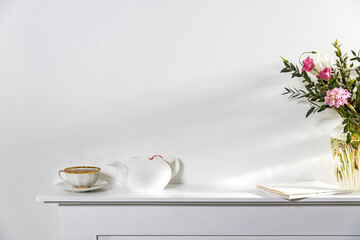 White on white. A cup of tea, a kettle, and a notebook stand on a white panel of an artificial fireplace.