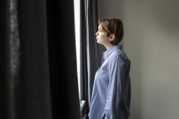 Fototapeta premium A pensive girl with a short hair nineteen years old in a white shirt with blue stripes looks out the window
