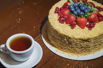 Close up multi-layered cake decorated with mixed berries. Blueberries, strawberries, redcurrant on small plate and cup of tea or coffee on brown wooden background. Copy space.