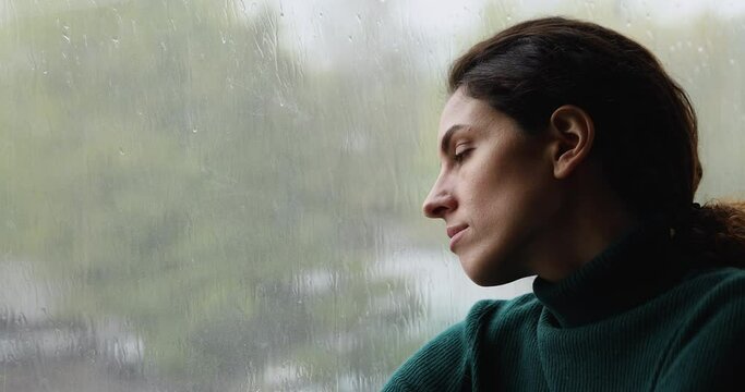 Close up head shot melancholic lost in thoughts young woman sitting near window, looking outside contemplating rainy gloomy weather, recollecting memories, dreaming or visualizing future alone indoors
