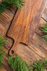 Cutting board on a wooden table background surrounded by pine branches, top view. Vertical.