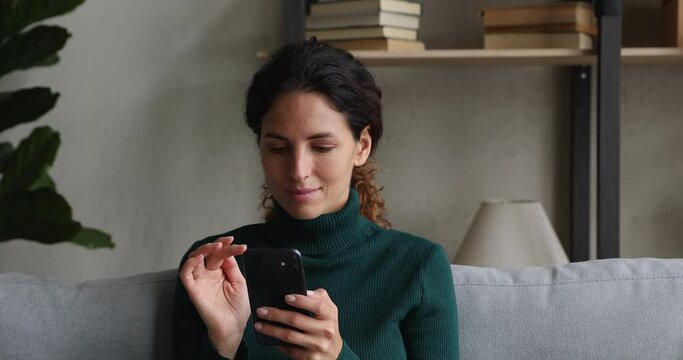 Happy millennial beautiful woman looking at mobile phone screen watching funny photo or video, enjoying pleasant distant communication in social network, web surfing or reading good news at home.