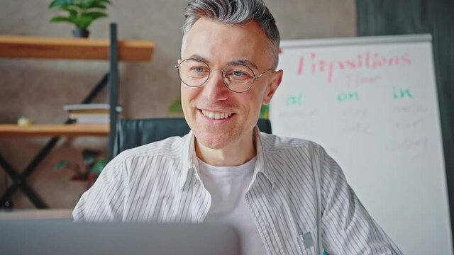 Positive middle aged tutor waving hand to laptop camera, video calling with student and explaining English rules online