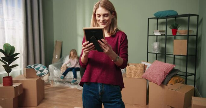Close up of cheerful young Caucasian beautiful woman unpacking carton boxes looking at photo frame with her husband moving into new apartment decorating home. Relocation concept. Settle in together