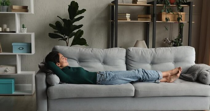 Full length happy millennial woman sleeping on comfortable couch in modern living room, breathing fresh conditioned humidifies air, meditating relaxing napping, enjoying peaceful weekend time.