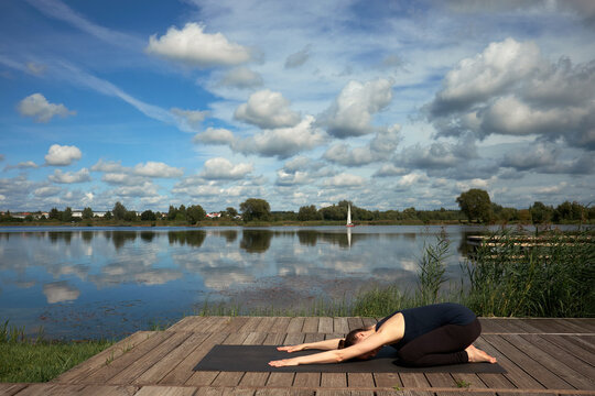 Slim young woman practicing yoga on the wooden pier near lake.