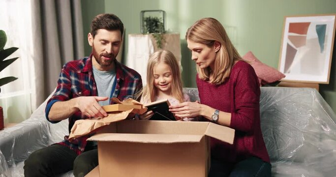 Joyful Caucasian happy cute parents sitting on sofa with small adorable Caucasian daughter kid opening big carton box unpacking stuff looking at photo frames in new apartment. Moving concept