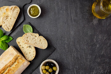 Fototapeta na wymiar Fresh italian ciabatta bread with herbs, olive oil, black and green olives, basil leaves and pesto sauce on dark background. Top view. Copy space.