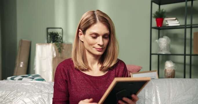 Camera approaching pretty cheerful young woman sitting on covered ofa in new apartment moving into new home and unpacking stuff opening carton box and holding photo frame in hands. Close up