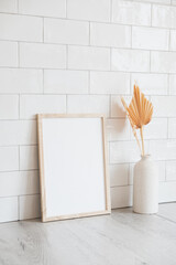 White vertical photo frame mockup on wooden table with dry flowers in vase. Scandinavian style home interior, neutral colors. . White tiles wall background.