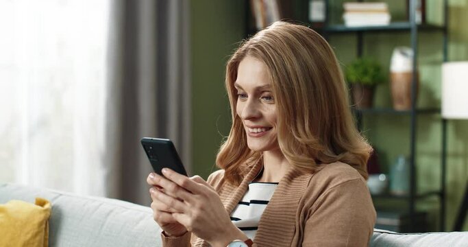 Close up of beautiful blonde woman sitting on sofa with smartphone in hands and laughing reading news online.