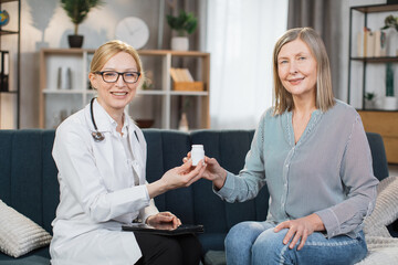 Experienced pleasant smiling woman doctor, giving bottle of pills to her sick female patient, prescribing treatment during home visit. Doctor and patient sitting on the couch at home.