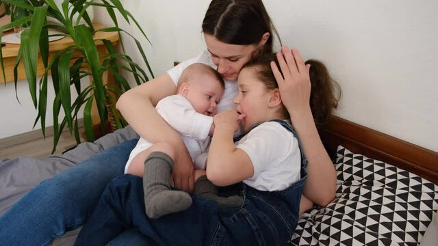 Caring mother hold lean to chest cute little infant toddler and preschooler daughter sit on bed, loving mom hug embrace two small sisters child, relax enjoy tender family moment. Childcare concept