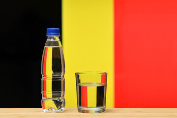 A bottle of clean drinking water and a glass stand on the table against the background of the flag of Belgium. A concept for the supply of clean drinking water in Belgium.