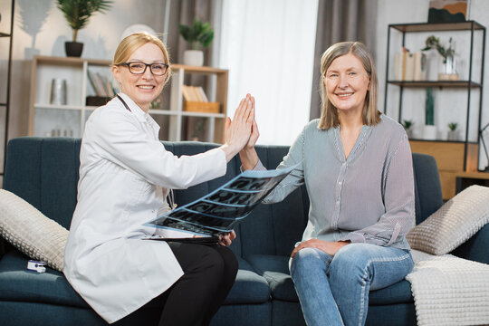 Cheerful positive blond woman doctor satisfied from successful treatment and result of x-ray scan of her retired female patient and giving high five each other during home visit