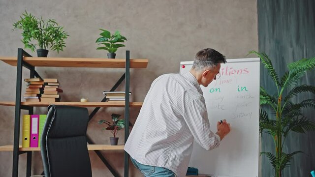 Positive middle aged man teacher explaining English rules to camera, writing examples om whiteboard at classroom