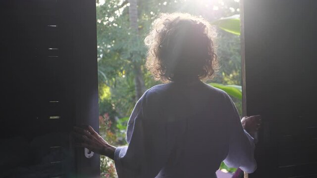Back view of woman opening a window and stretching. Sunny morning in Bali