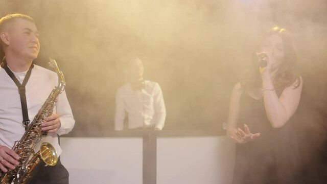 Bearded DJ standing at the mixer controller, composing a new mix the party in nightclub. Musical group of singer vocalist woman, saxophonist sax, dj man playing song performing on concert with lights.