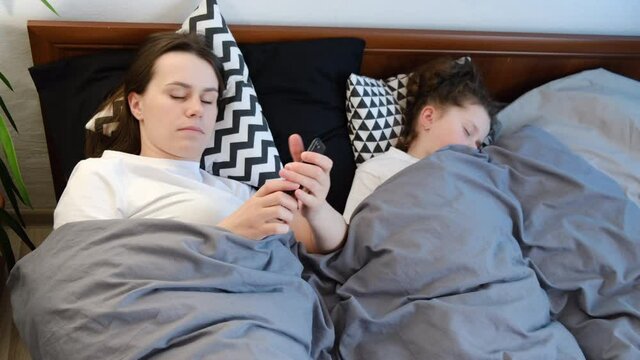 Close up of young caucasian mother using phone browsing net website lying on cozy bed ay home near sleeping cute little daughter, beautiful brunette mom put away mobile phone at night ready to sleep